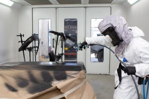 Person wearing full overalls and breathing apparatus in a vehicle bodyshop paint spraying booth, spraying car parts with black paint